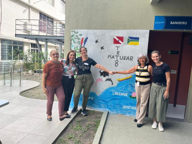 Fotografia em ambiente aberto. Na foto está presente 5 mulheres, todas estão sorrindo na frente de uma parede que acabou de ser pintada— Foto: ASCOM8