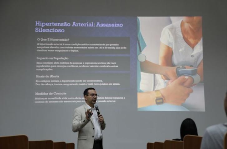 Descrição de imagem: Fotografia em ambiente fechado, centralizado, está o cardiologista, Rodrigo Souza, falando ao microfone.