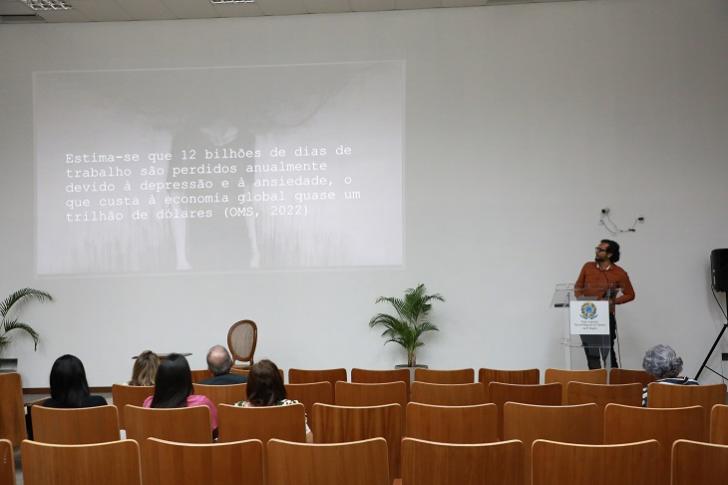 Fotografia colorida do auditório da EJUD, na foto está 6 pessoas sentadas assistindo a palestra e o palestrante Dr. Eric Alvarenga no palco.