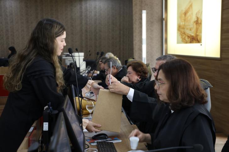 Foto colorida. O desembargador Itamar Junior deposita o voto na urna. Próximo a ele os demais desembargadores na bancada do tribunal pleno.