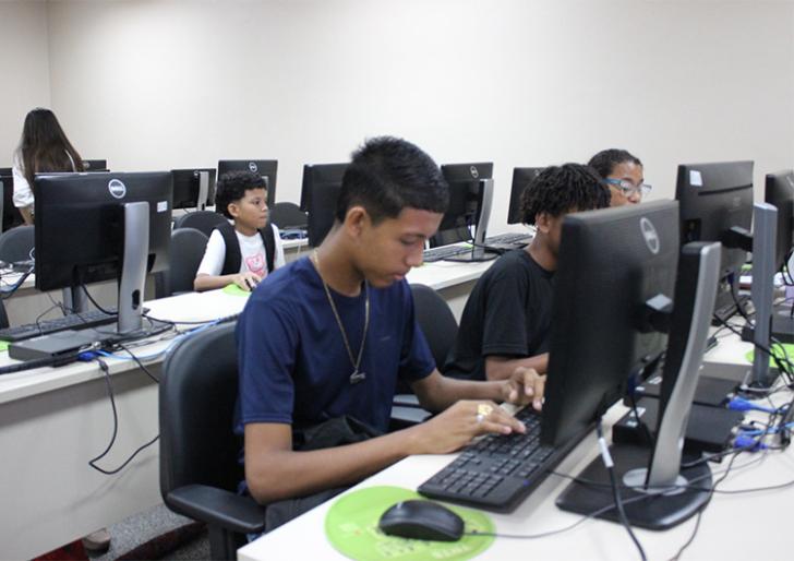 Descrição de imagem: Fotografia contendo algumas crianças e adolescentes mexendo em computadores.