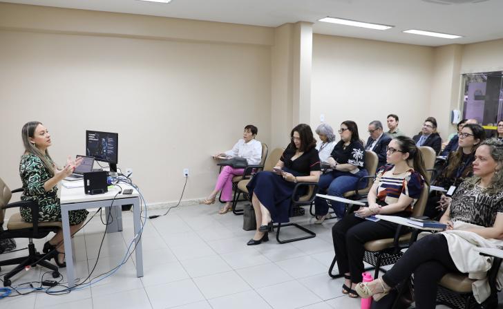 Foto colorida mostra momento de aula de precedentes. A professora está sentada à mesa e fala aos alunos.