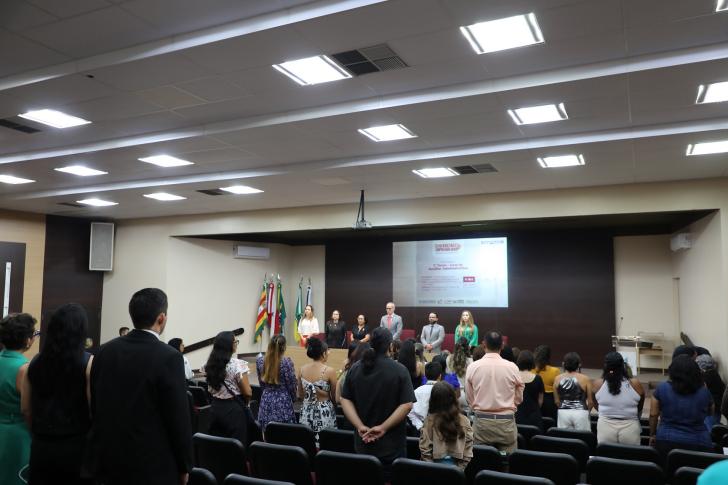 Foto da cerimônia de formatura, com magistrados, convidados e formandas de pé no momento do hino nacional.