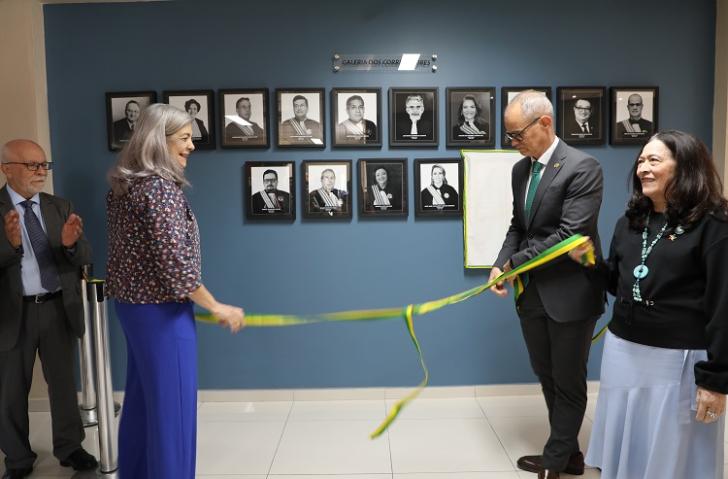 Fotografia em ambiente fechado que reúne um homem e duas mulheres, descerrando a fita de inauguração de uma exposição