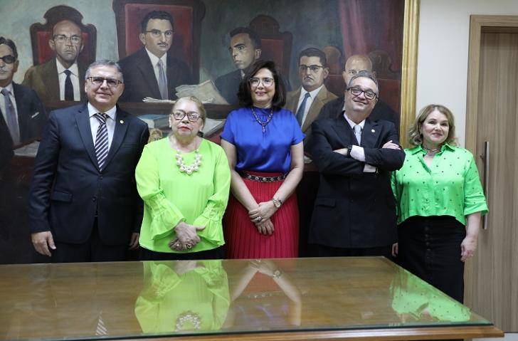 Fotografia em ambiente fechado, nela, estão cinco pessoas de pé atrás de uma mesa.  