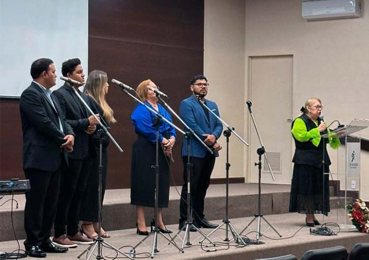 Fotografia em ambiente fechado que reúne um grupo musical formado por homens e mulheres e uma mulher branca em destaque. 