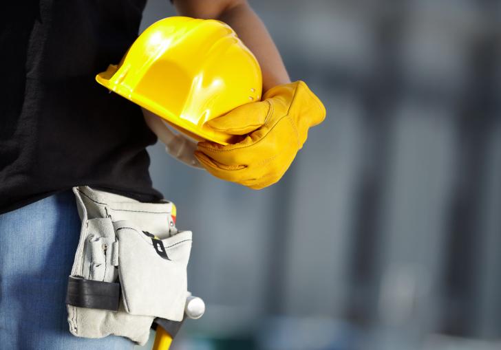 A imagem mostra uma pessoa segurando um capacete de segurança amarelo com uma das mãos, que está protegida por uma luva amarela de trabalho. 