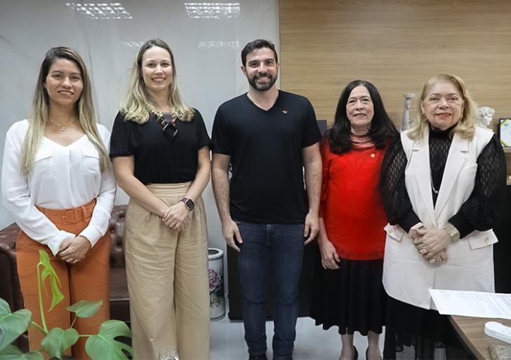 Descrição de imagem: Fotografia em ambiente fechado, nela, estão cinco pessoas, quatro mulheres e um homem. Todos estão de pé e sorriem para a foto. 