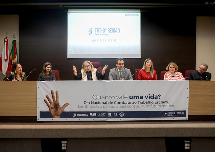 Descrição de imagem: Fotografia em ambiente fechado. Centralizadas, estão sete pessoas sentadas no palco do auditório do TRT-8.  