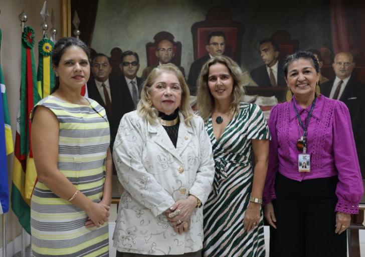 Descrição de imagem: Fotografia em ambiente fechado, nela, estão quatro mulheres de pé e sorrindo para a foto.