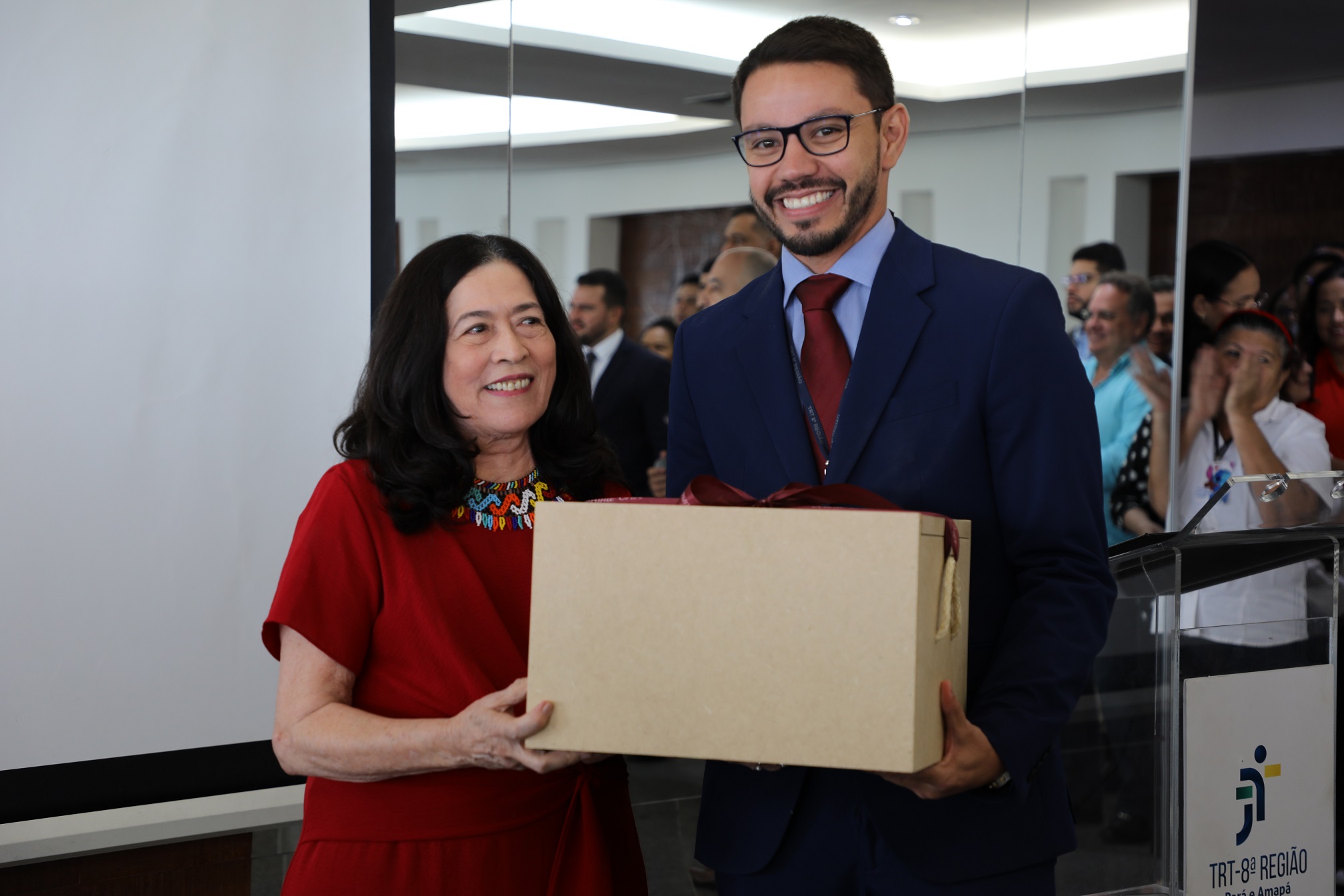 Foto colorida da desembargadora Zuíla Lima Dutra, ao lado do servitor Heitor Junior, durante entrega de uam caixa de presentes para a corregedora regional.