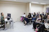 Foto colorida mostra momento de aula de precedentes. A professora está sentada à mesa e fala aos alunos.