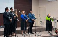 Fotografia em ambiente fechado que reúne um grupo musical formado por homens e mulheres e uma mulher branca em destaque. 